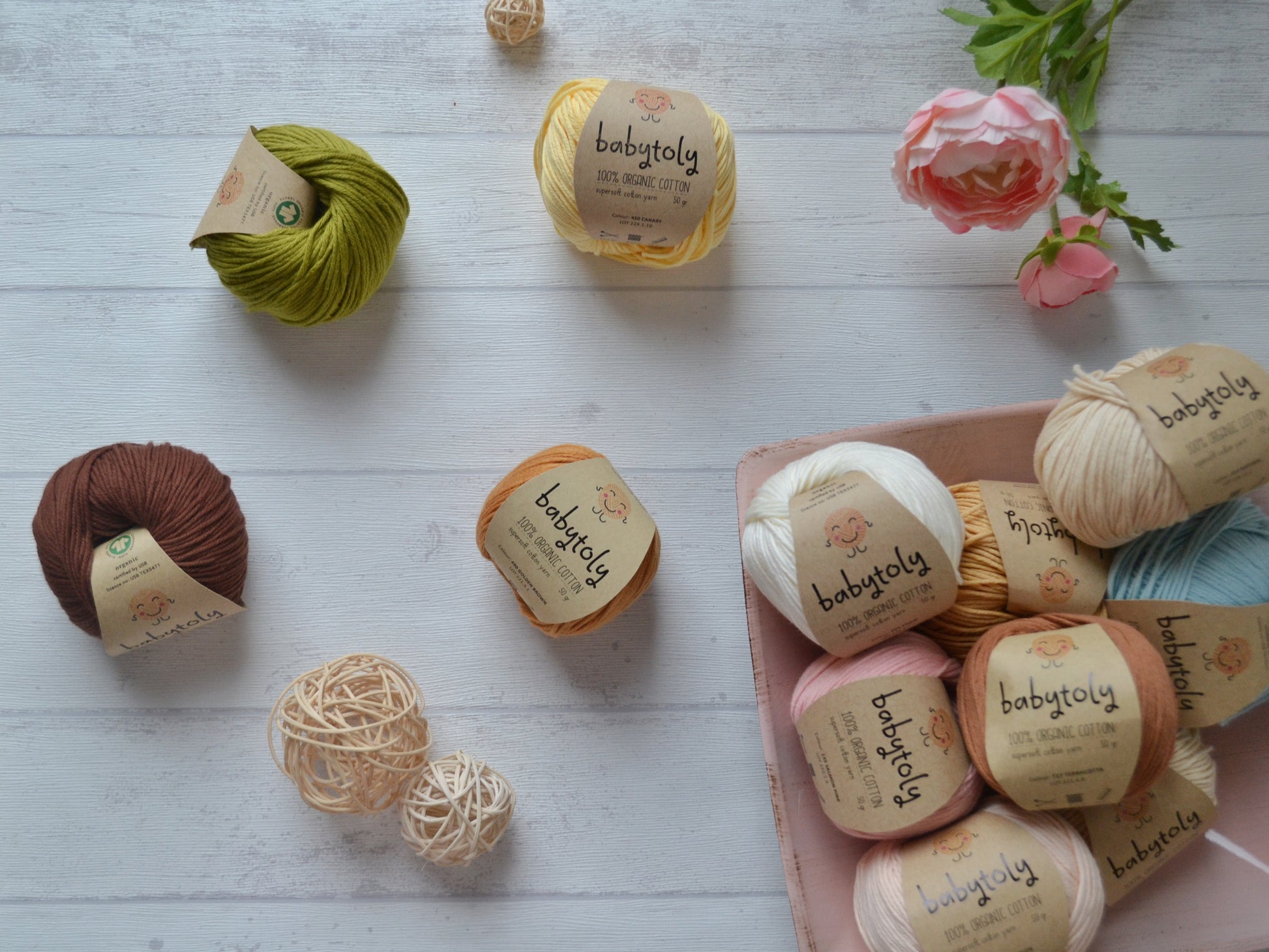 Rechts unten eine Schale voller Babytoly Organic Cotton. Links davon sind 4 Knäuel wild verteilt mit verschiedenen Farben Babytoly Organic Cotton. Oben rechts ist eine Kunstblume abgebildet. Unten Links sind zwei Cremefarbene runde Dekoelemente abgebildet. 