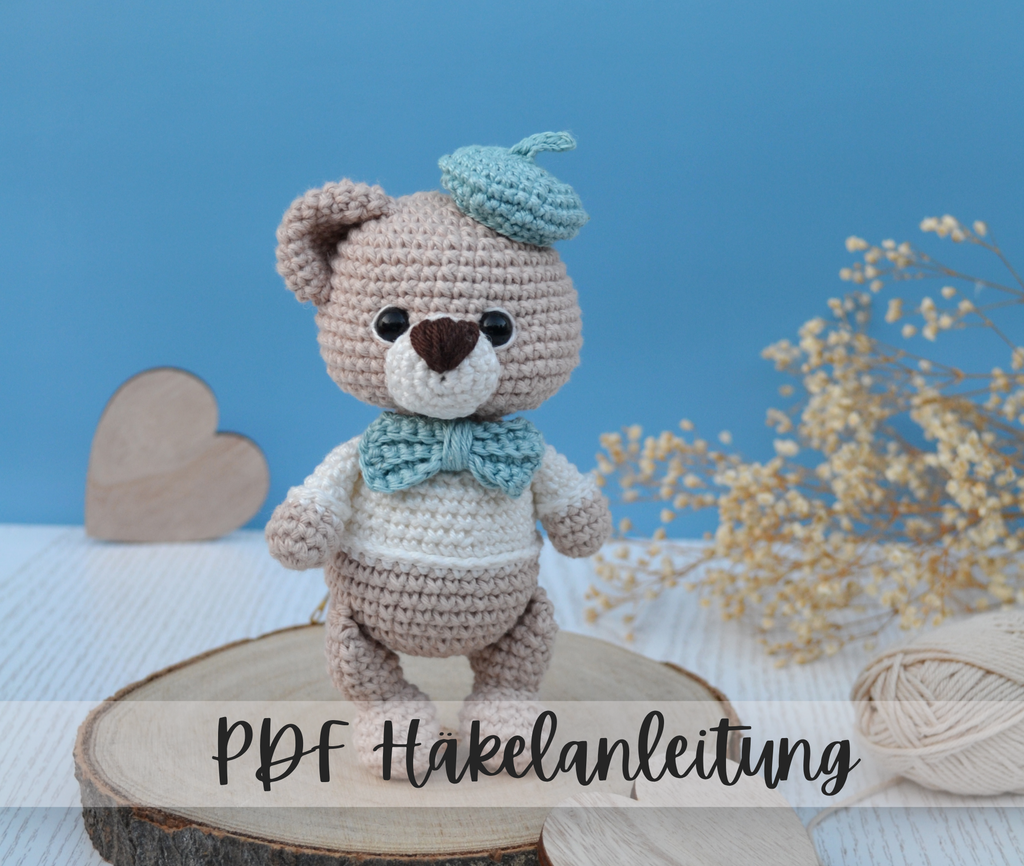 Ein Amigurumi Bär mit blauer Baskenmütze und Schleife am Hals der niedlich in die Kamera schaut. 