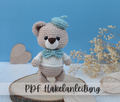 Ein Amigurumi Bär mit blauer Baskenmütze und Schleife am Hals der niedlich in die Kamera schaut. 