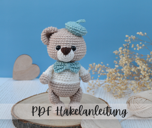 Ein Amigurumi Bär mit blauer Baskenmütze und Schleife am Hals der niedlich in die Kamera schaut. 