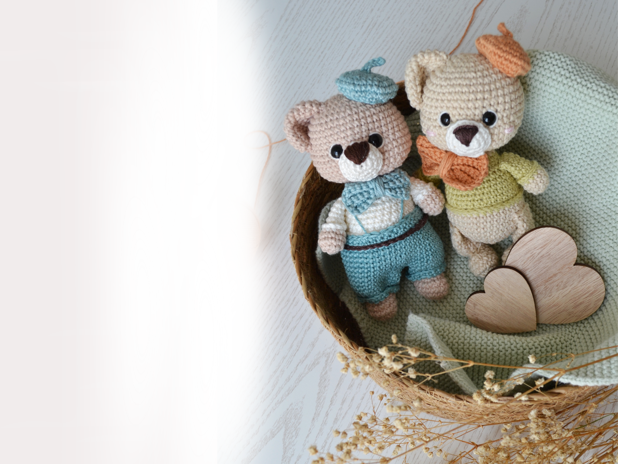 Zwei gehäkelte Bären mit Mütze auf dem Kopf und Schleife am Hals, sitzen in einem Korb aus Seegras mit einem Stricktuch und zwei Herzen aus Holz. 